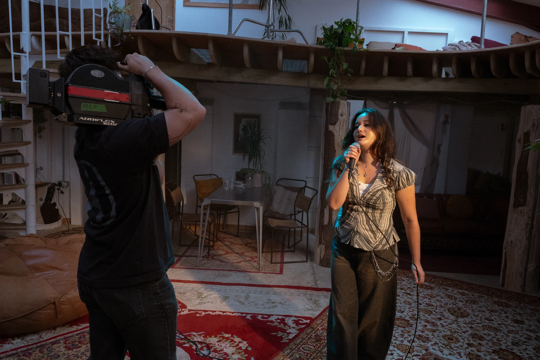 A camera operator films Clarissa Mae singing into a microphone with a shoulder-mounted film camera on the bed crumbs set.