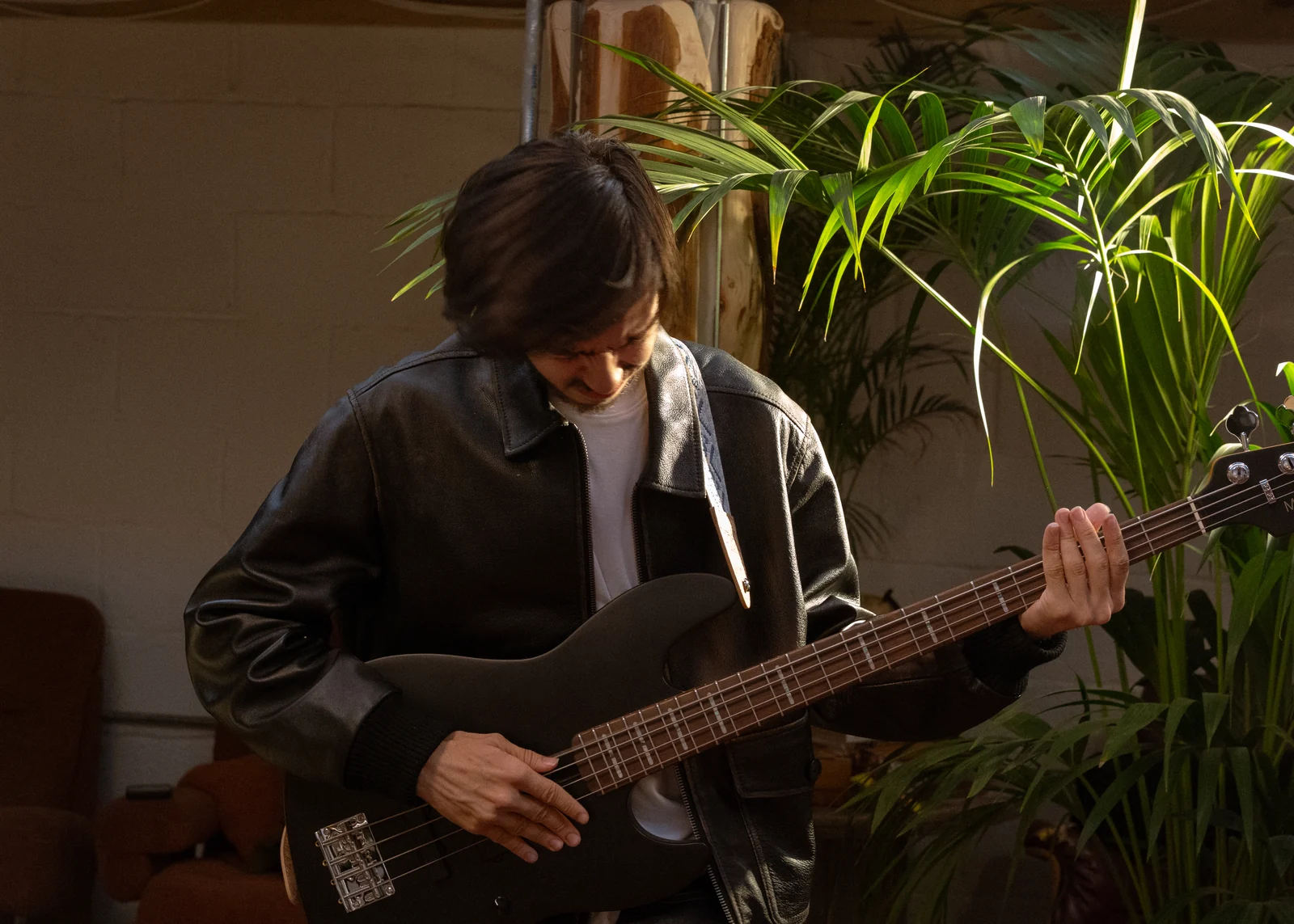 KDYN in a black leather jacket playing a black five-string bass guitar beside plants on the bed crumbs video set.