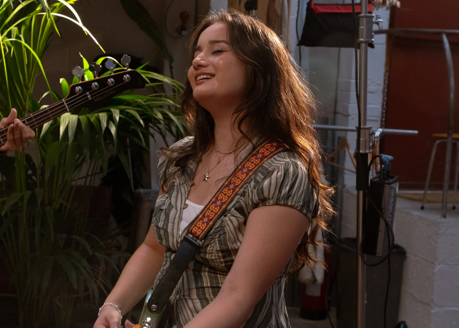 Clarissa Mae smiling with an electric guitar during filming of the bed crumbs music video, surrounded by studio plants.